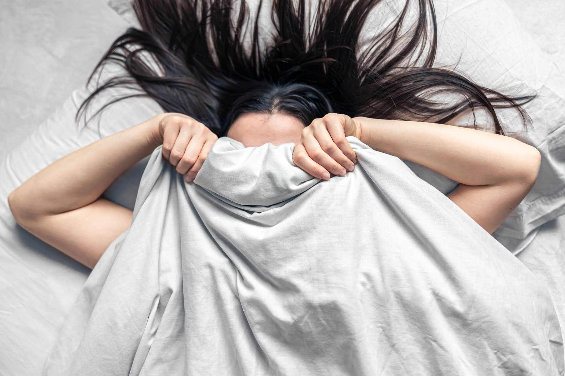 Woman lying in bed pulling the blanket over her face, representing hypersomnia, where sleeping more never feels like enough.